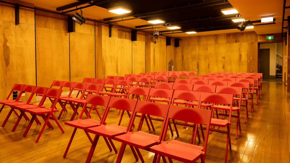 A room prepared for a presentation. Rows of red folding chairs are arranged on a wooden floor facing the back of the room, with wood paneling. The black ceiling features square lights and stage lights. A doorway with a green exit sign is situated on the right side of the room.