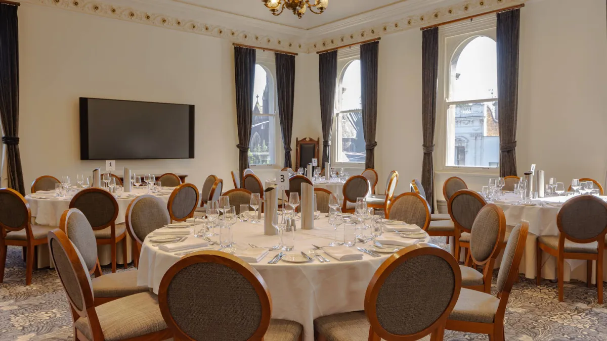 A formal dining room, featuring round tables with white tablecloths, silverware, glassware. Each table is encircled by wooden chairs with grey upholstered seats. A large television screen is mounted on the wall between two windows with dark grey curtains. The room is adorned with decorative crown molding and a patterned carpet, with a chandelier hanging from the ceiling.