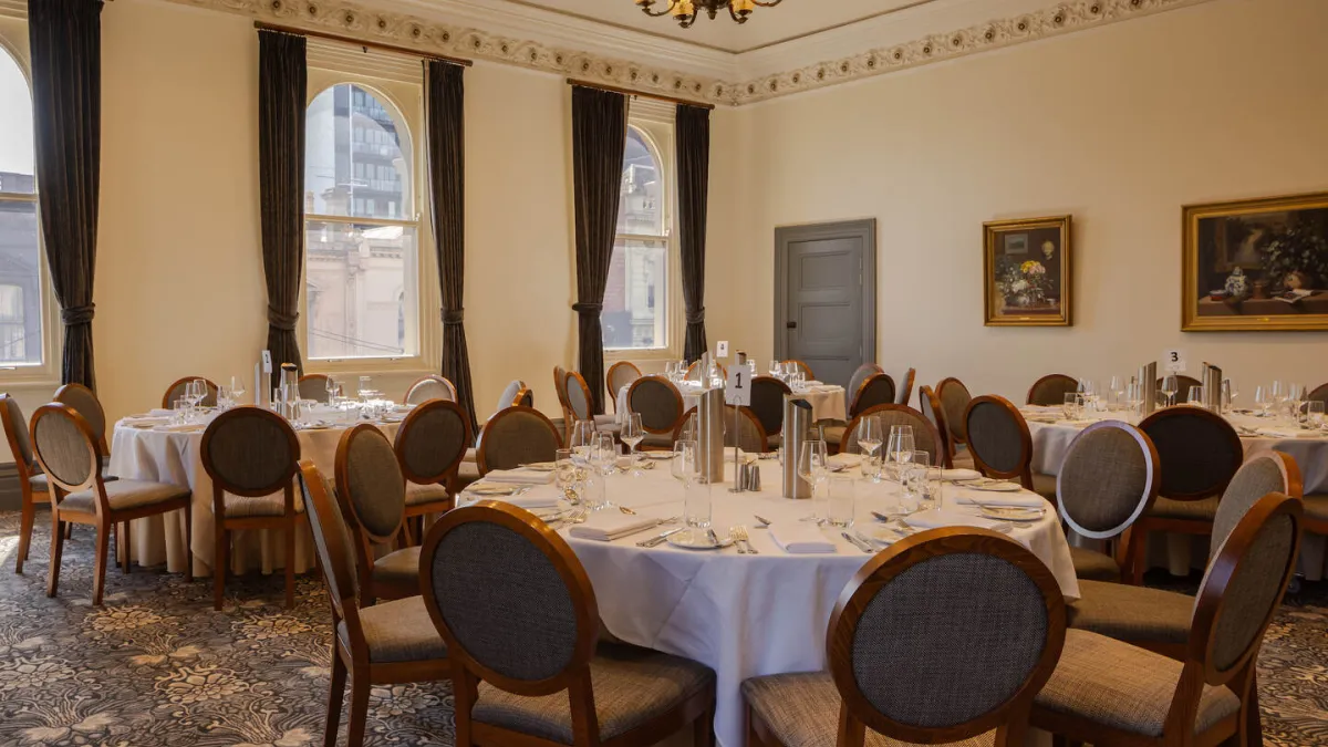 A room set for a formal event with round tables dressed in white tablecloths, surrounded by chairs, and equipped with plates, silverware, and glassware. Tall windows are adorned with brown drapes, and light-colored walls display paintings. The room features a chandelier and patterned carpet.