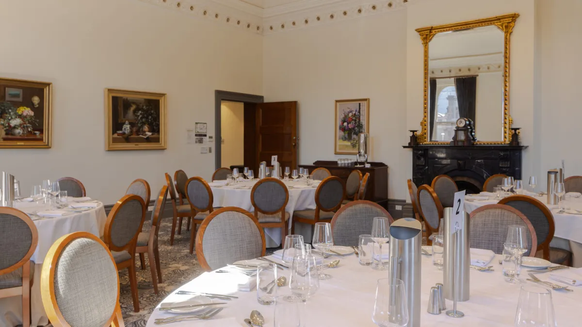 A banquet hall set with round tables draped in white tablecloths and formal place settings. Artwork hangs on the  walls, and a large mirror is above a fireplace. A chandelier is visible on the ceiling