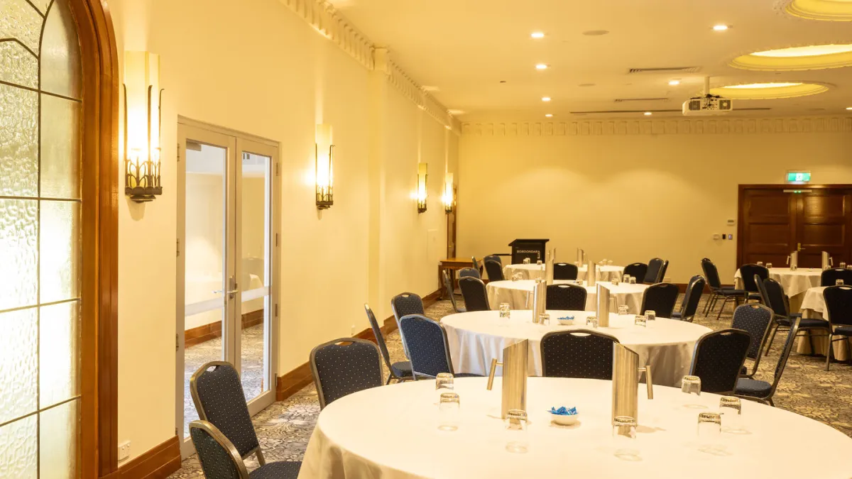 A room with round tables covered with white tablecloths, each set with glasses and a water pitcher, and surrounded by blue chairs. The room has decorative lighting on the walls, and a podium in the background. A large window is on the left of the room.
