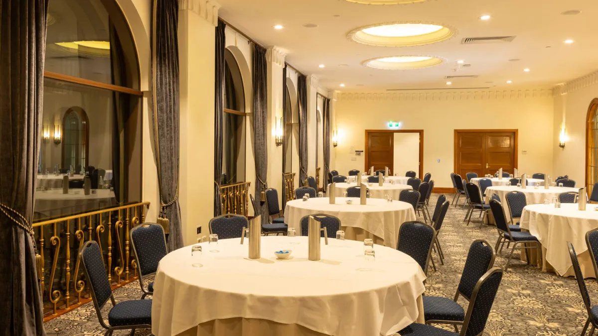 A conference room is set up with round tables covered in white cloths, each surrounded by chairs with blue patterned upholstery. There are large, arched windows with dark curtains along one side of the room. The ceiling features recessed lighting and a projector. The floor is covered with a patterned carpet.