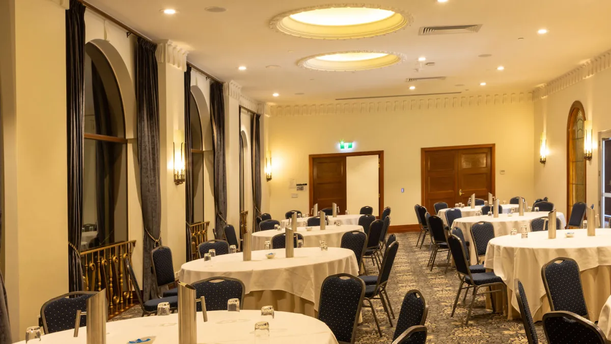 A large ballroom space set up for an event. Round tables with white linens and blue chairs are arranged throughout the room. Tall arched windows are on the left with heavy grey curtains. The ceiling has decorative molding and circular light fixtures. A projector is mounted on the ceiling. Two sets of wooden doors are at the far end of the room, one marked as an exit.
