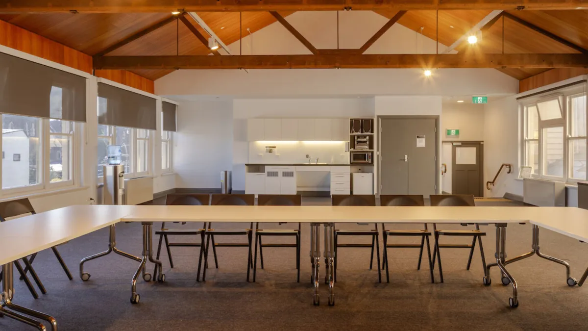 A meeting room featuring a U-shaped white table, dark chairs, a carpeted floor, and a kitchenette in the background. The ceiling has exposed wooden beams. Windows with shades provide natural light.