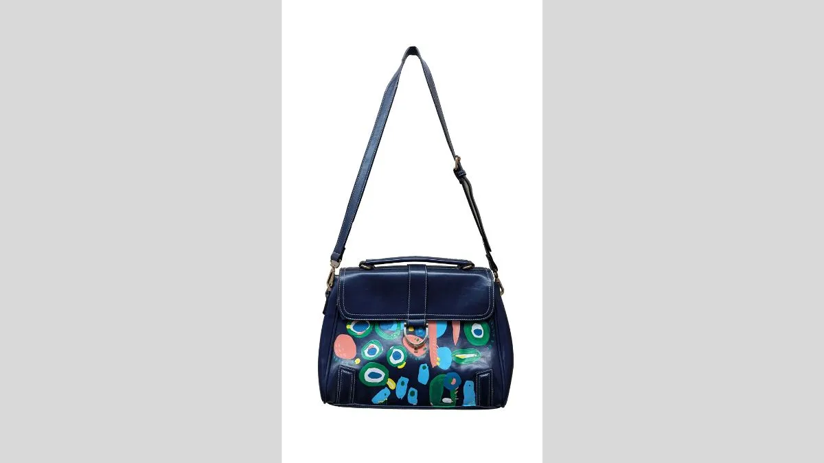 A blue bag with a shoulder strap. Body of bag is painted with graphic shapes in pink, green, white, blue and yellow. 