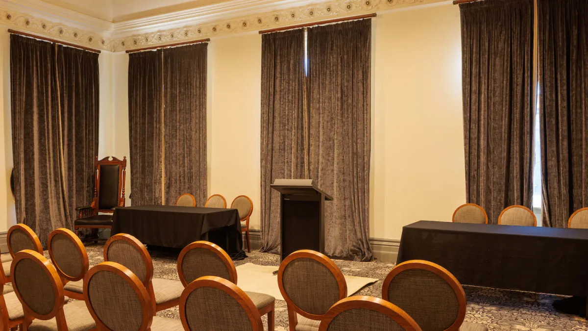 A room arranged for a presentation. Rows of chairs face a podium and tables with black cloth. Windows are covered with dark brown curtains. An ornate chandelier hangs from the ceiling.