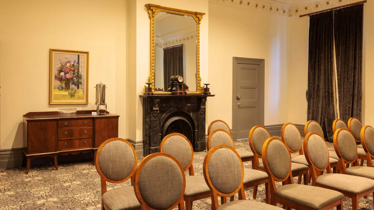 A room arranged for a presentation, with rows of chairs facing forward. The room has a buffet table, floral painting, a coffee urn,  a black marble fireplace, a mirror. A doorway is to the right of the fireplace. The room has decorative molding around the ceiling and patterned carpet.