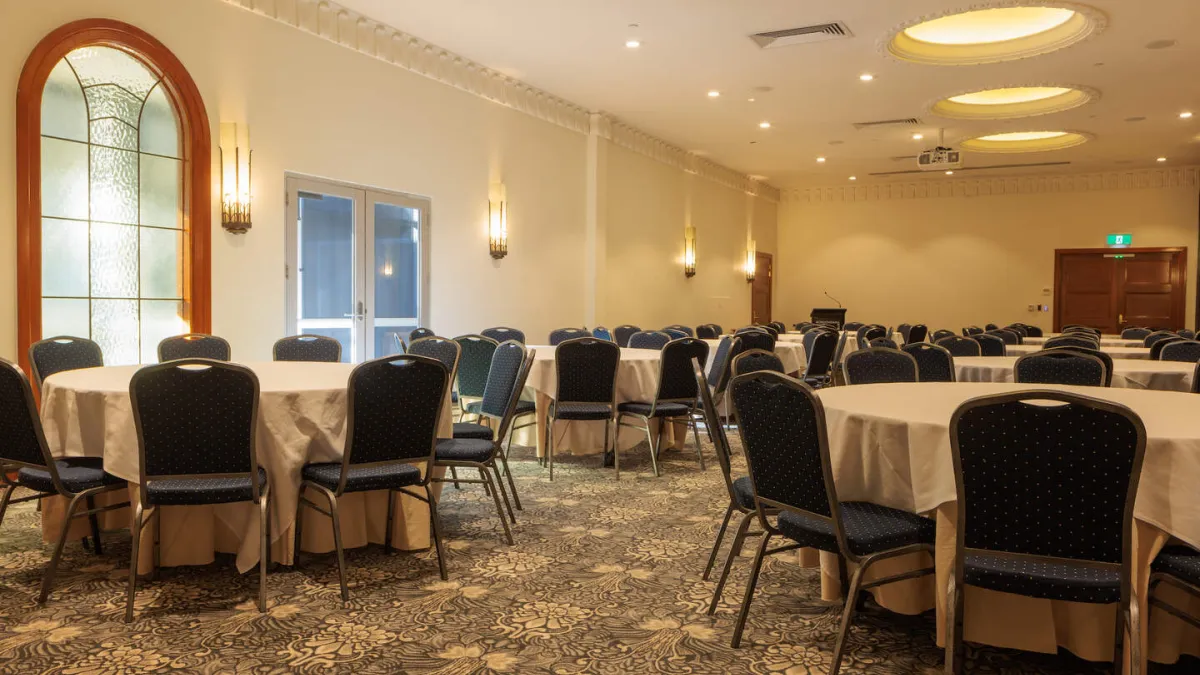 A spacious banquet hall is arranged for an event with round tables adorned with white tablecloths, encircled by chairs. The room boasts ornate ceiling details and lighting fixtures, a sizable window, and patterned carpeting. A projector is mounted on the ceiling at the rear of the hall.