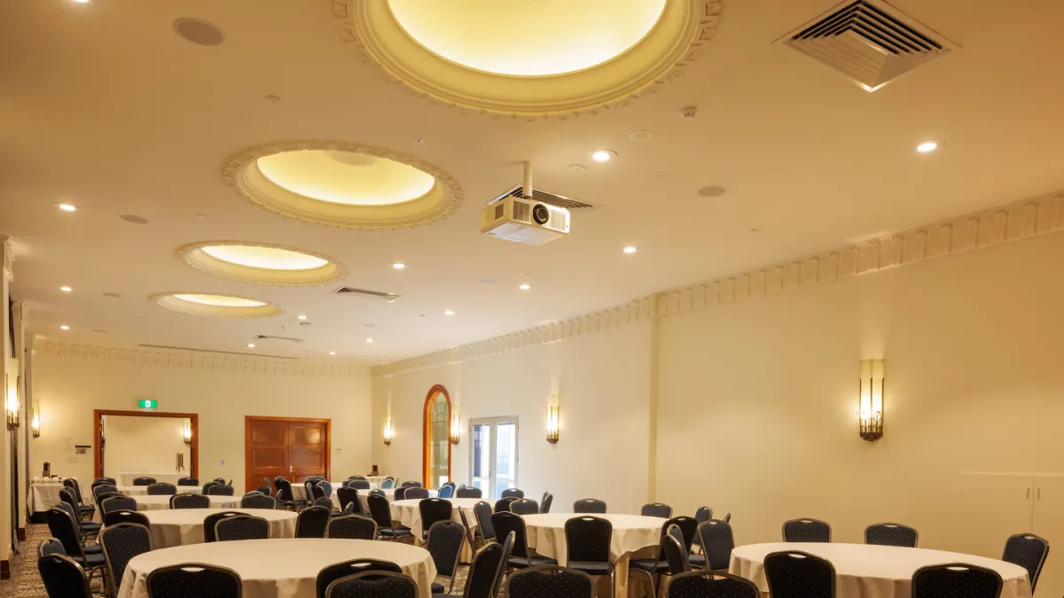 A large, formal banquet room with round tables covered in white cloths, surrounded by black chairs. The room has a decorated ceiling with recessed lighting and wall sconces.