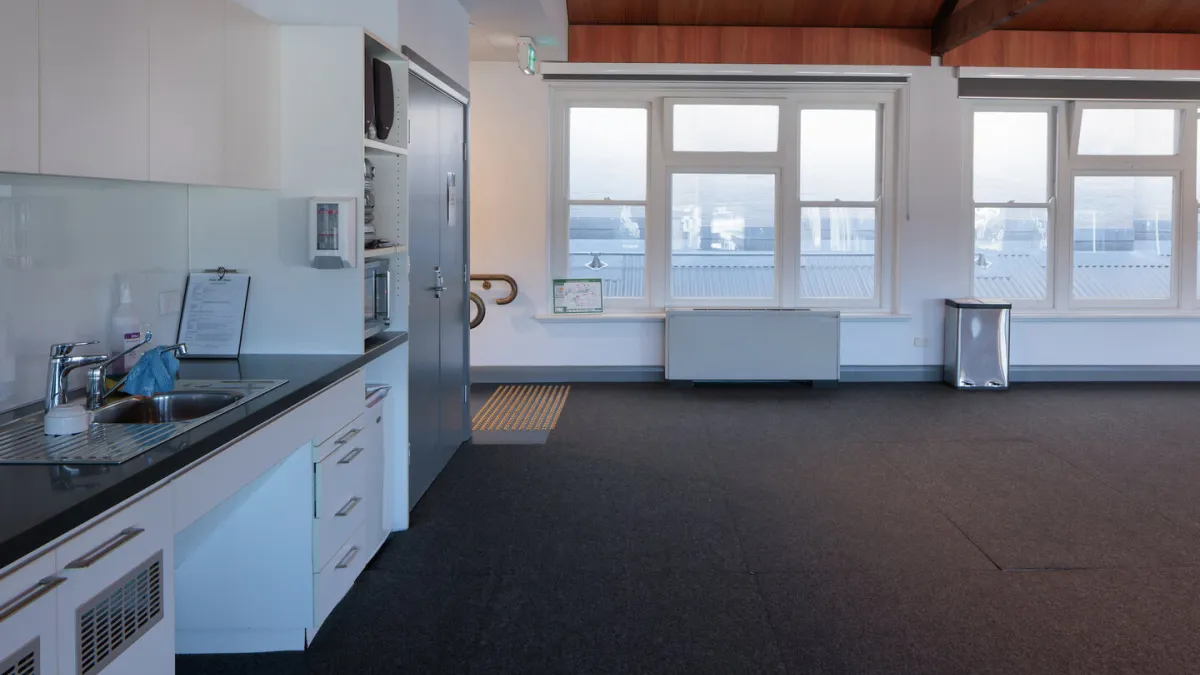 A room with a kitchenette, large windows, and dark gray carpeting. A sink, cabinets, and microwave are on the left. Two large windows are on the right with a radiator and trash can.