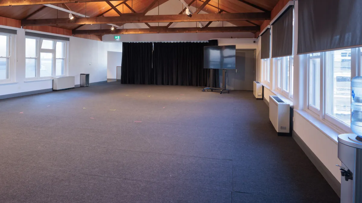 An event space featuring a large open floor plan, natural light from multiple windows, a stage area with black curtains, and a large display screen on wheels.