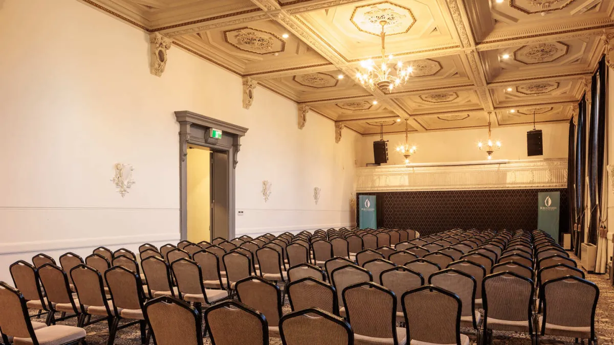 A spacious room is prepared for a presentation, with rows of chairs facing a stage adorned with two vertical banners. The room features an ornate, vintage ceiling and chandeliers. An open doorway is visible on the left.