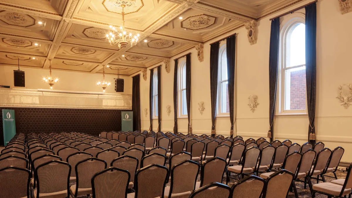 An interior shot of a formal event space with rows of chairs facing a stage. The room features ornate ceiling details, chandeliers, and tall windows with dark curtains. The stage has speakers on either side and banners.