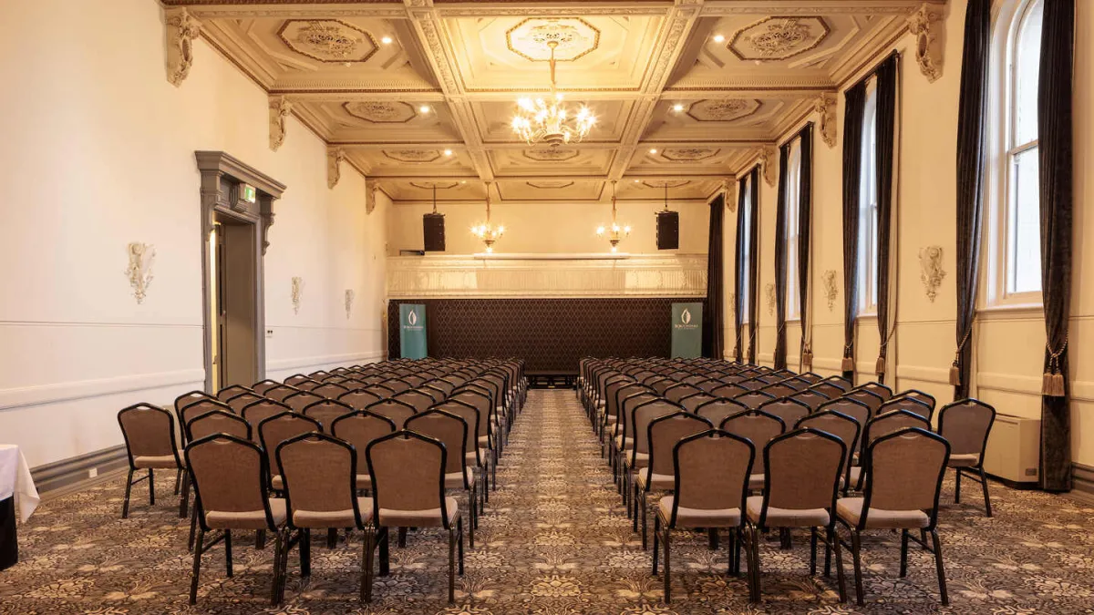 An interior view of a large room set up for an event. Rows of chairs face the back of the room, where a raised platform is centered.  The ceiling is ornately decorated with square recessed panels and gold trim and chandeliers  Tall windows line one side of the room, draped with dark curtains. The floor is covered with a patterned carpet.