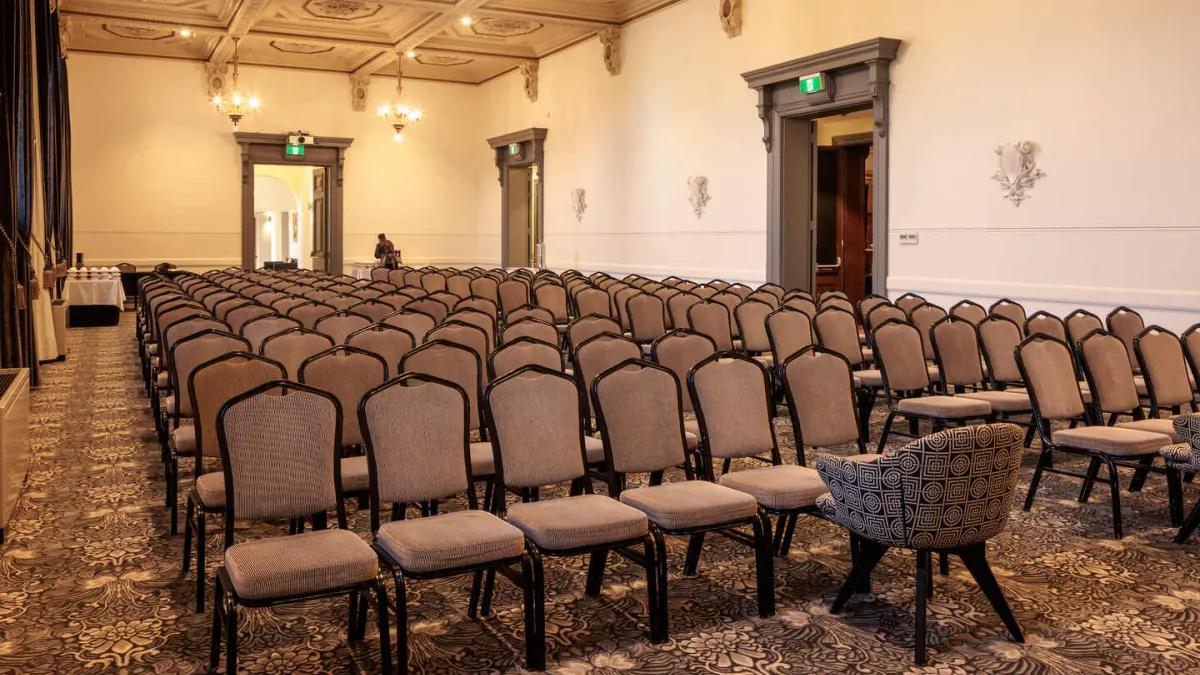 A lecture hall or presentation room, with rows of chairs facing the front. The room has a decorated ceiling and doors with exit signs. There is a patterned carpet.