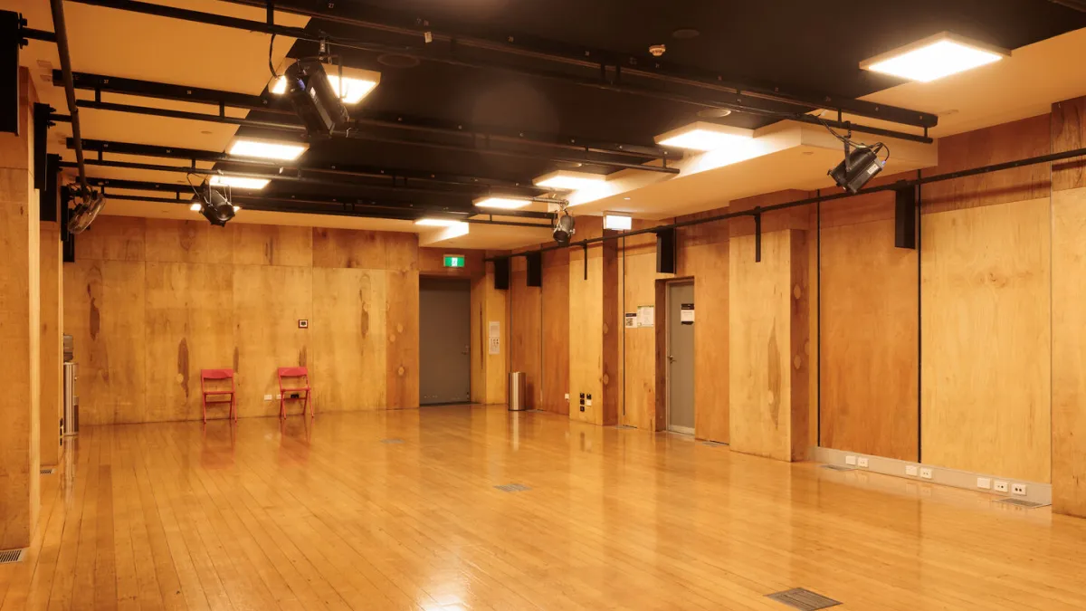 Interior view of a dance studio or theater rehearsal space with wooden floors and walls, overhead lighting and rigging, and two red chairs.