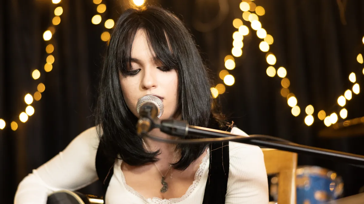 A dark haired woman with a guitar sings into a mic with fairy lights behind her