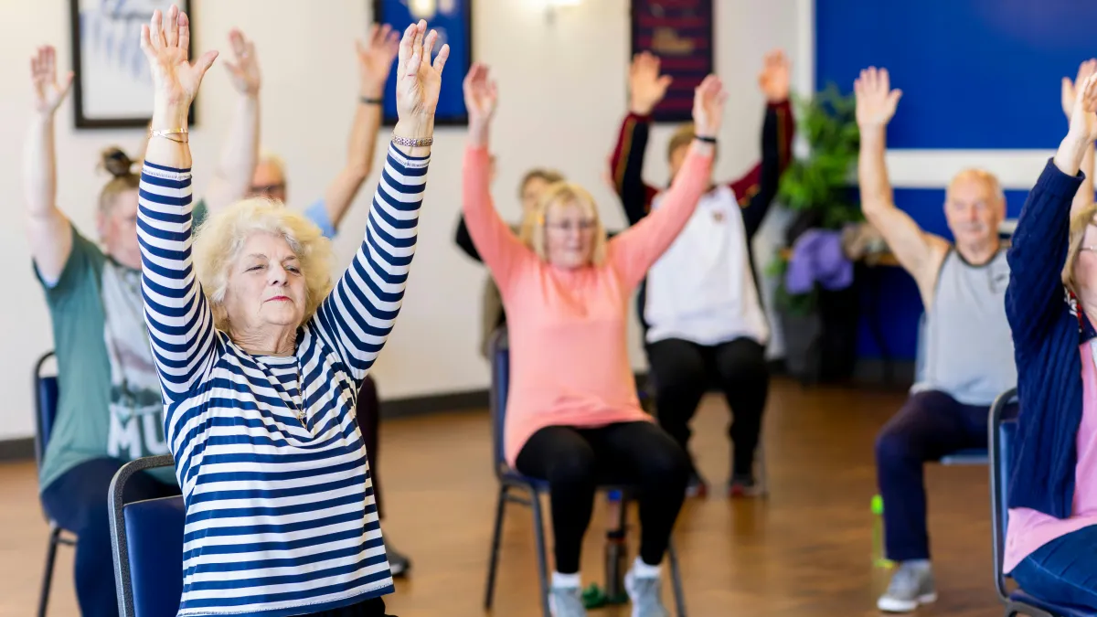 A group of older adults are sitting on chairs with their hands in the air. 