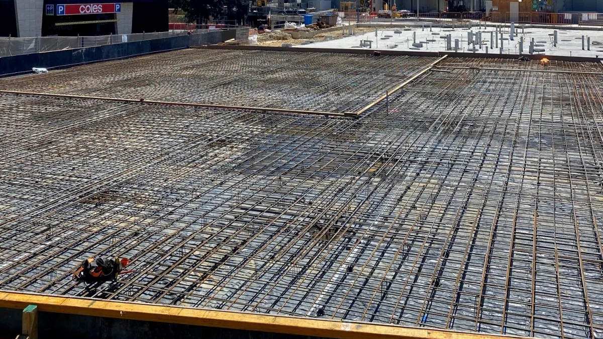 In the foreground, a dense grid of reinforcing steel (rebar) is laid out, ready for concrete pouring. Behind the rebar, there's evidence of ongoing construction activities, including concrete foundations, exposed framework, and equipment. In the background, there's a larger structure under construction, with its concrete walls partially completed.