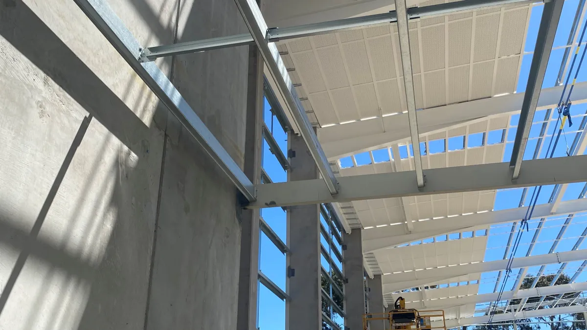 A low-angle perspective of a building under construction. Steel beams frame the structure against a blue sky. Concrete walls stand to the left, and a worker is visible on a yellow lift in the distance, working on the roof.