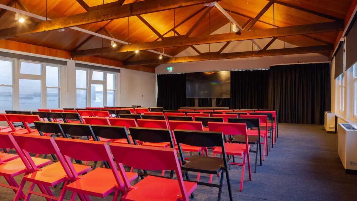 Rows of folding chairs are set up in a room for a presentation. A large screen hangs on the wall behind black curtains. The room has a high wooden ceiling with exposed beams, and windows line the left and right walls.