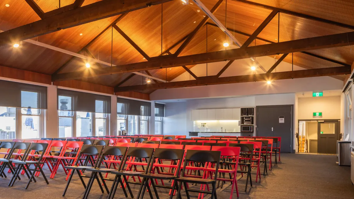 a large hall with a high, peaked wooden ceiling. Rows of folding chairs looking forward. Windows with closed blinds are on the left. A white kitchenette area is at the back, next to a gray door. Exit signs are illuminated above a door on the right.