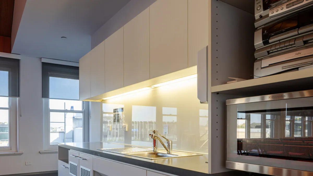A modern kitchen space featuring a stainless steel sink, white cabinets, a microwave, and a vintage stereo system. Two windows with roller shades provide a view of a cityscape.