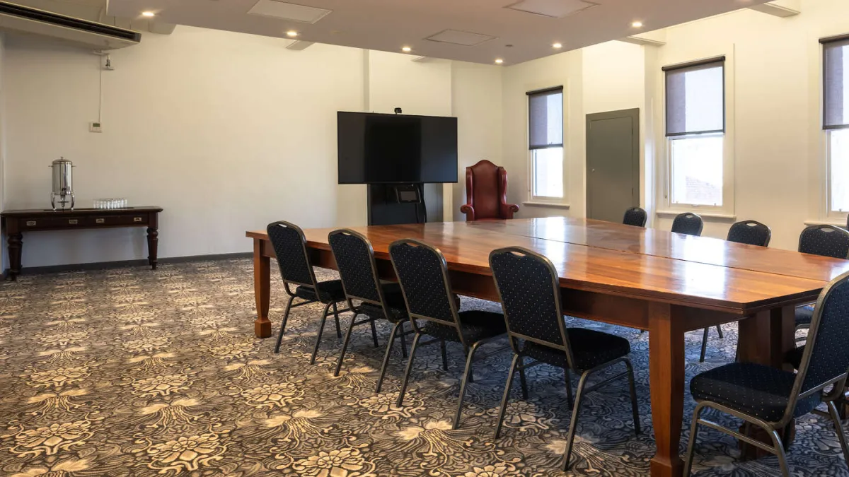 A conference room featuring a long wooden table, black chairs, a large flat screen television, and a water dispenser on a table. The room has windows with blinds and a patterned carpet.