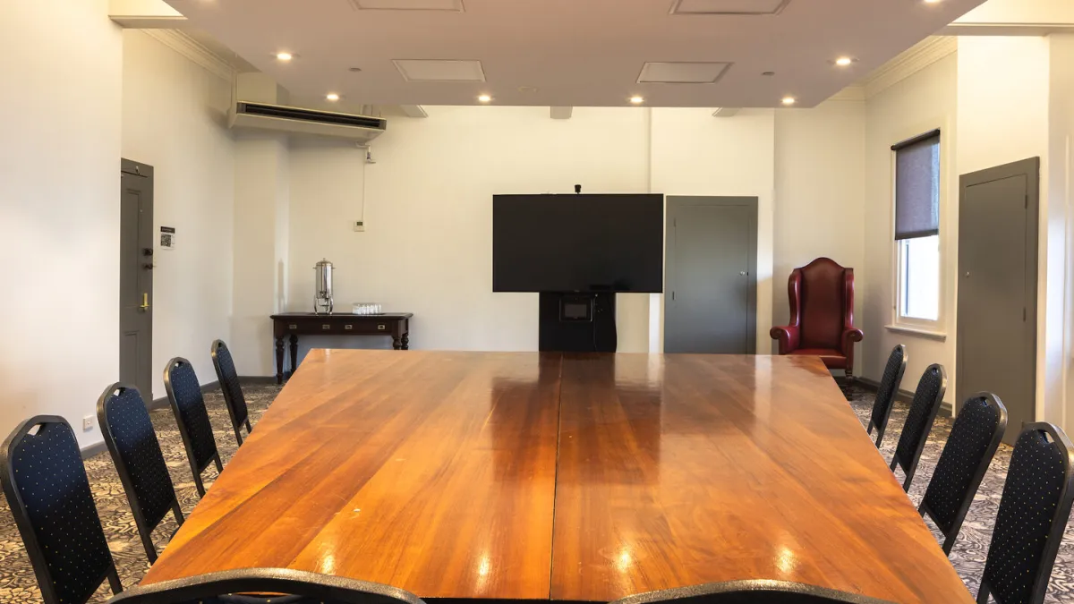 A conference room is furnished with a A conference room is furnished with a long wooden table, chairs, and a large television screen. There is a water cooler on a table, and a red chair. There is one window. The floor is carpeted with a white floral pattern on a dark background. 