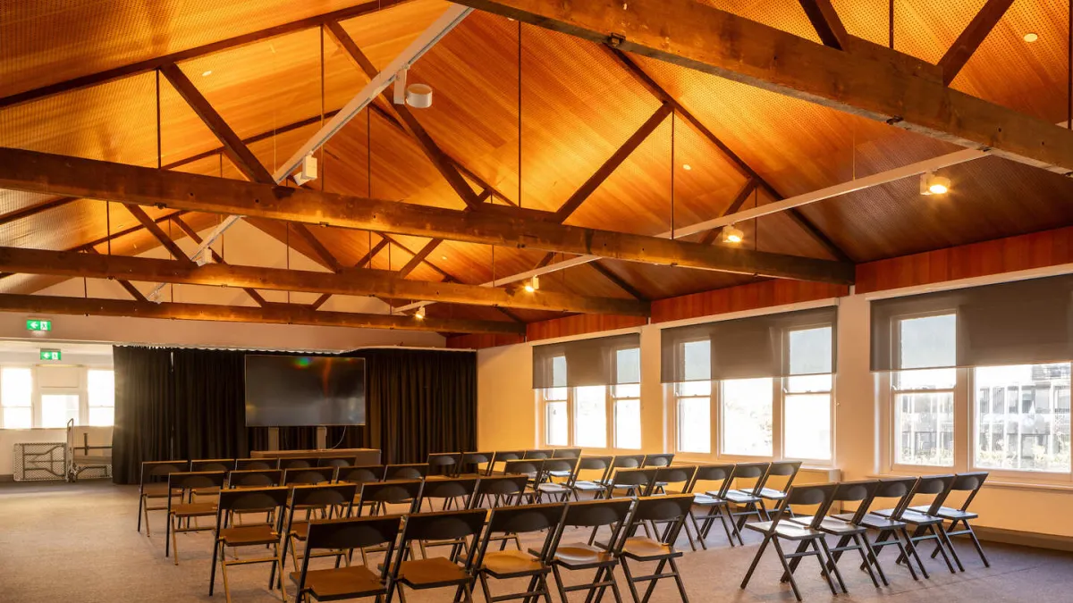 A room is arranged for a presentation with rows of folding chairs facing a screen and black curtains. The room features a high, wood-paneled, gabled ceiling with exposed beams. Natural light enters through a row of windows on the right.