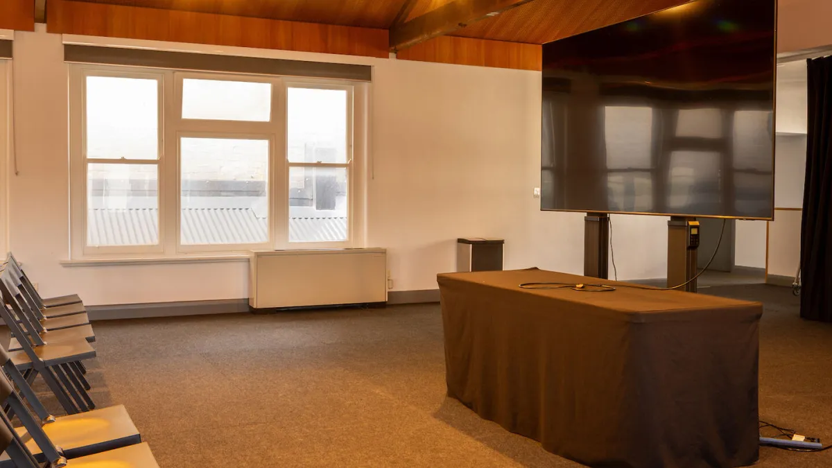 A room set up for a presentation. Folding chairs line the left side of the frame, facing a large television screen in the middle of the room, behind a draped table. A window to the left of the screen lets in natural light. The room has gray carpeting and a wood ceiling.