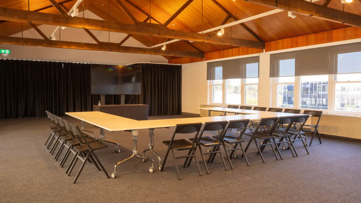 A spacious meeting room featuring a high, exposed wooden ceiling. A large U-shaped table is in the centre, surrounded by black chairs. A prominent screen is set against black curtains. Windows with gray blinds line one wall. The floor is carpeted in gray.