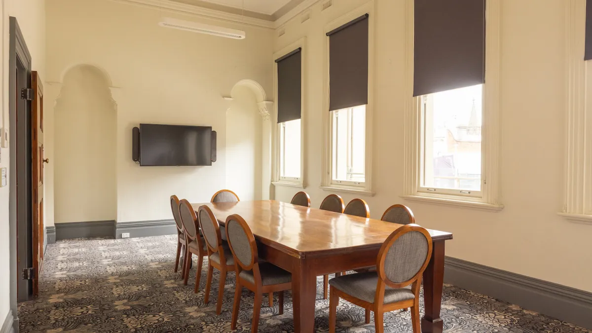 A meeting room featuring a long rectangular wooden table with 10 chairs. A TV sits on the far wall. There are tall windows and patterned carpet. 