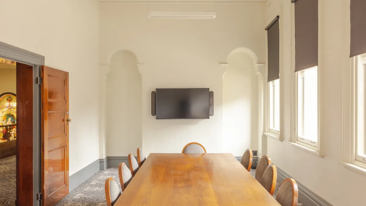A conference room featuring a long rectangular wooden table down the centre surrounded by 10 chairs. There is a TV mounted on the far wall. A door is to the left. 