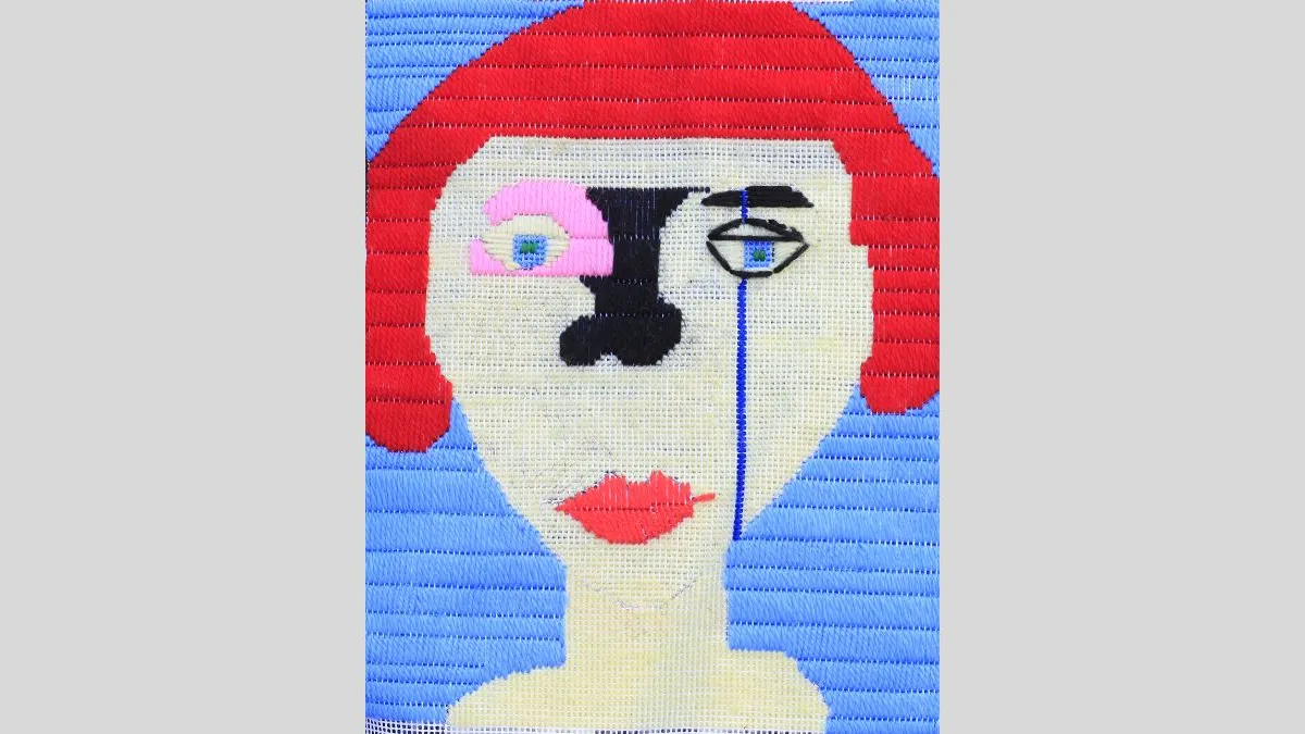 Image of a woven textile artwork featuring ga stylised portrait. The figure has read hair, light skin and red lips. The right eye is blue and outlined with black stitching and a long blue line extending down the face to the chin. The left eye is pink. The background is blue. 