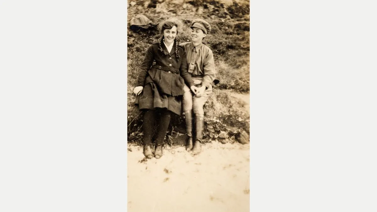 A woman in a chauffeur outfit holds hands with a woman whose hair is in plaits