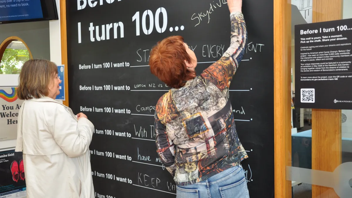 Two women writing their Before I turn 100 statements on a blackboard