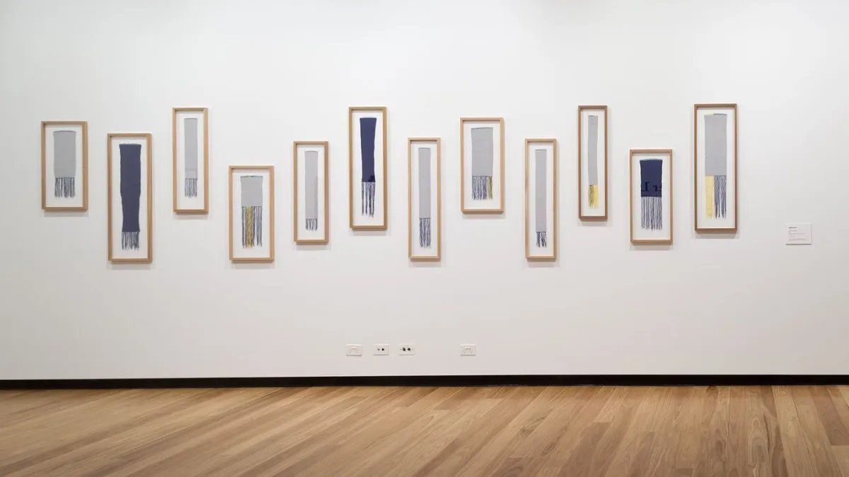 A photograph of an installation of framed small tapestries on a white wall.