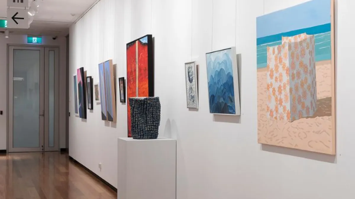 The image shows a long, well-lit gallery space with wooden flooring and white walls adorned with various artworks. The artworks include colourful abstract paintings, geometric compositions, and landscape scenes, all framed or mounted on the walls. The perspective extends down the hallway.