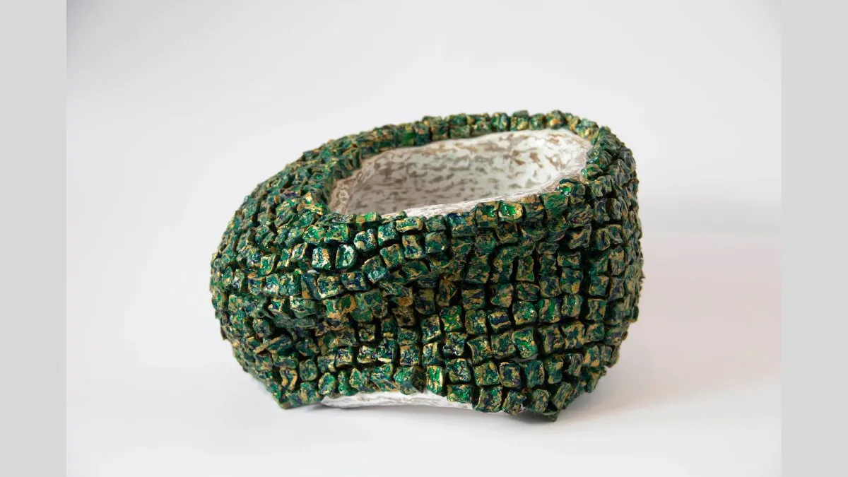Small sculptural vessel with an uneven, textured exterior made of raised cube-like forms. The cubes are painted with green and gold, and the interior is smooth and pale cream. The sculpture is photographed against a white background.