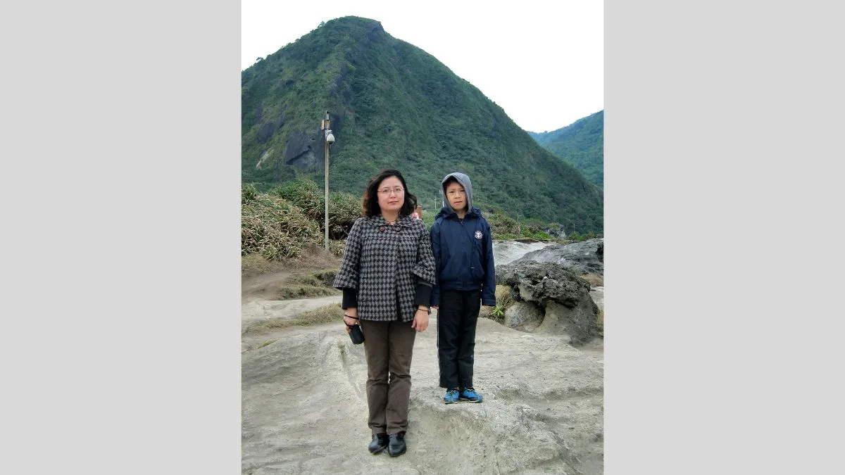 A mother and son stand together in front of a mountain. The son stands on a perch to be the same height as his mother. 