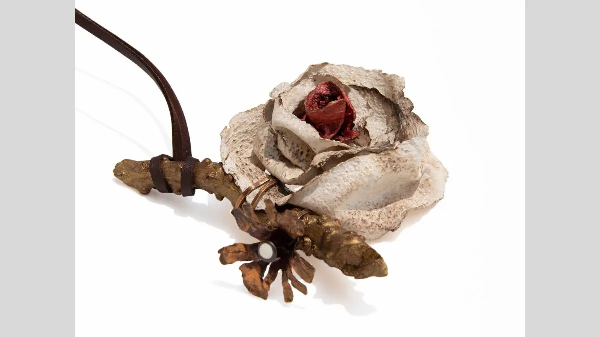 Small sculptural necklace featuring a pale, stone-like rose form with a dark red centre attached to a metal stem and mechanical-looking elements, set against a white background.