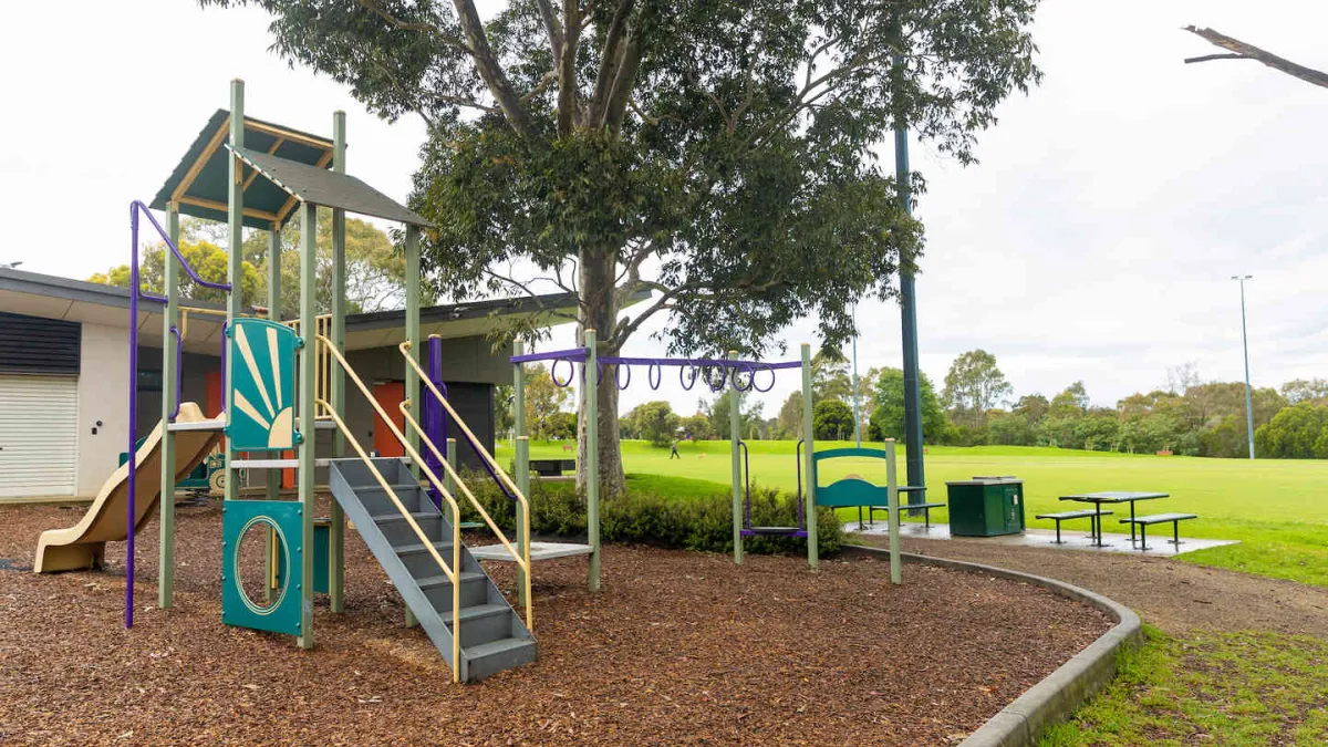 A playground featuring a slide, with a park lawn, picnic table and barbecue in the background