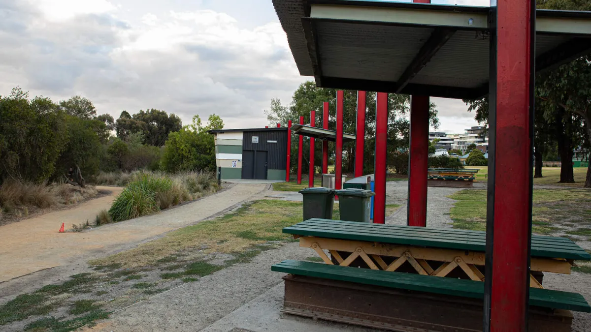 Modern park shelter with red support columns and a flat roof. Timber picnic tables are underneath the shelter, with waste bins nearby. A concrete path leads to building.