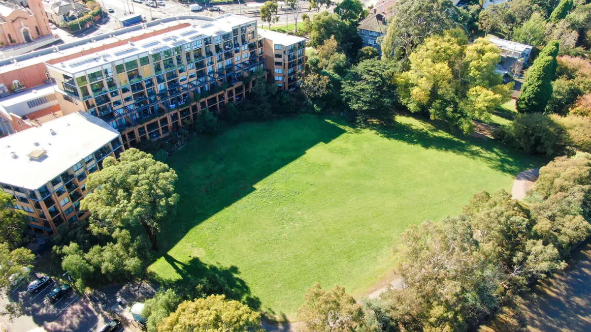 Birds eye view of a large open space park bordered by trees and a tall building. 