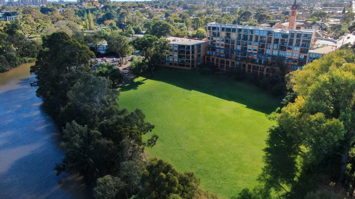 Birds eye view of a large square park space next to a river. The park is surrounded by trees and a apartment building