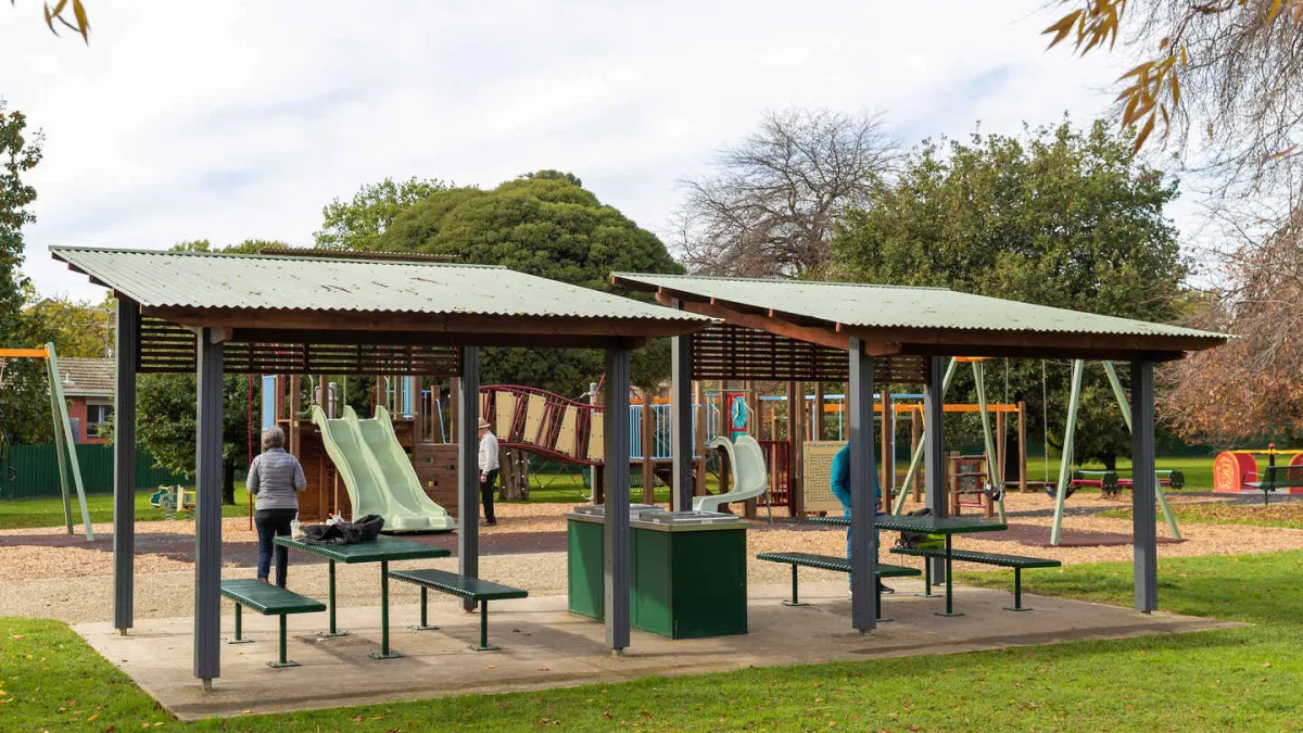 2 sheltered picnic shelters with a barbeque in between. the background is a children's playground
