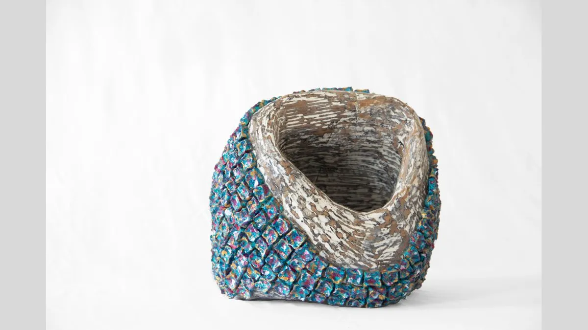 Small sculptural vessel with an asymmetrical opening, featuring a textured exterior of raised cube-like forms. The cubes are painted blue with pink highlights. The inside surface is smooth and painted in a creamy white colour, pattered with dark thin streaks. The sculpture is photographed against a white background.