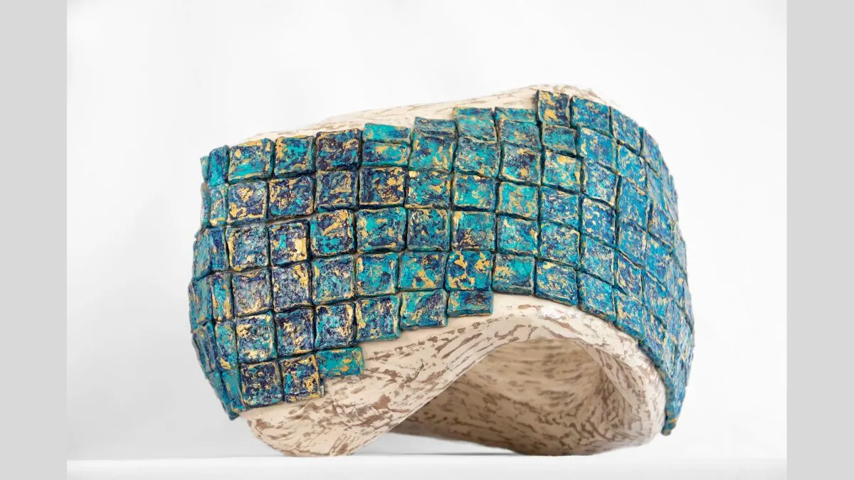 Small sculptural vessel with a curved, asymmetrical form, featuring a textured exterior of raised square tiles. The tiles are painted turquoise and gold. The interior of the sculpture is pale cream and marbled. The sculpture is photographed against a white background.