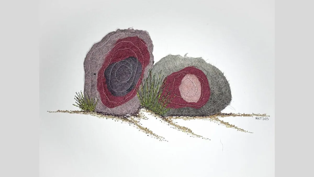 Photo of a mixed media textile artwork on white paper depicting two, rock-like forms. Each form is composed of layered and stitched pieces in muted tones of maroon, grey, black, and purple, creating circular patterns. Between and around the rock shapes, green and brown threads and textured fibres simulate tufts of grass and soil, extending slightly from the surface. 